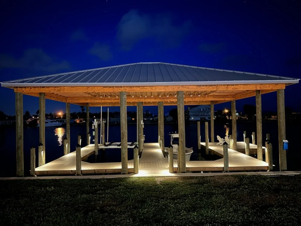 Night view of a wooden boat dock with a metal roof, illuminated by lights underneath, on a calm body of water with houses in the background.