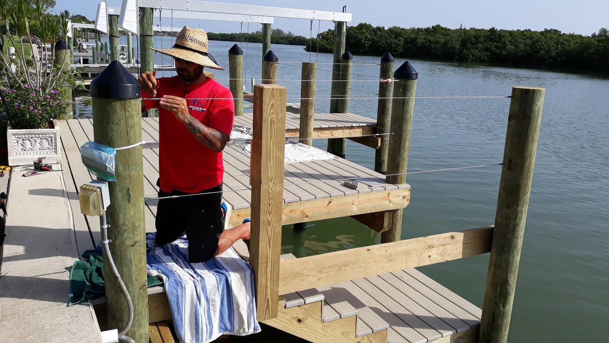 A man installing or repairing a dock on a body of water. He is kneeling on a towel, wearing a straw hat and a red t-shirt, with tools around him.