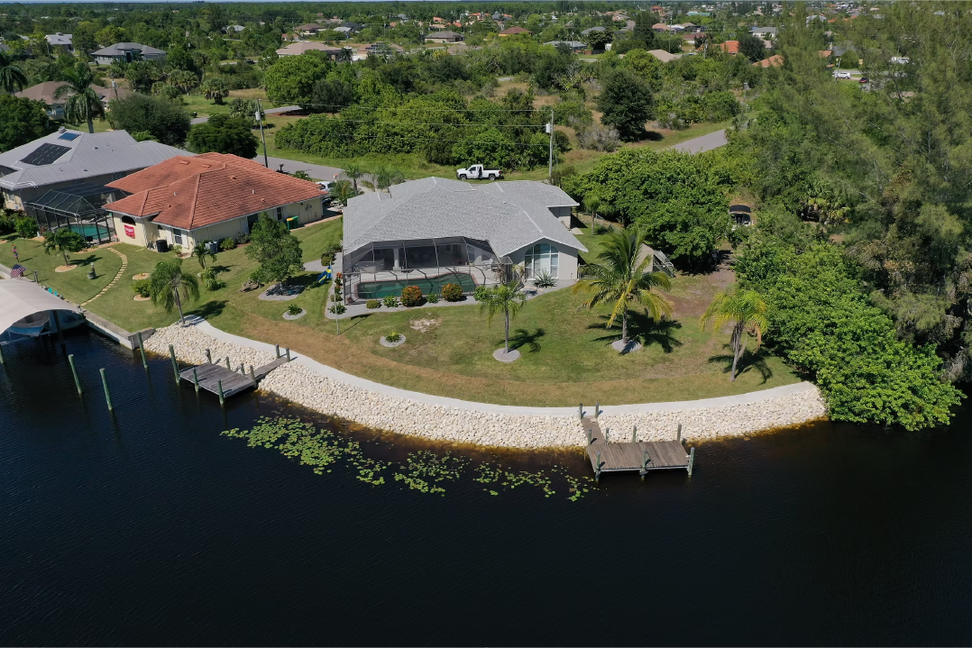 Aerial view of a waterfront house with a pool, palm trees, and a dock along a body of water, with neighboring houses and lush greenery in the background.