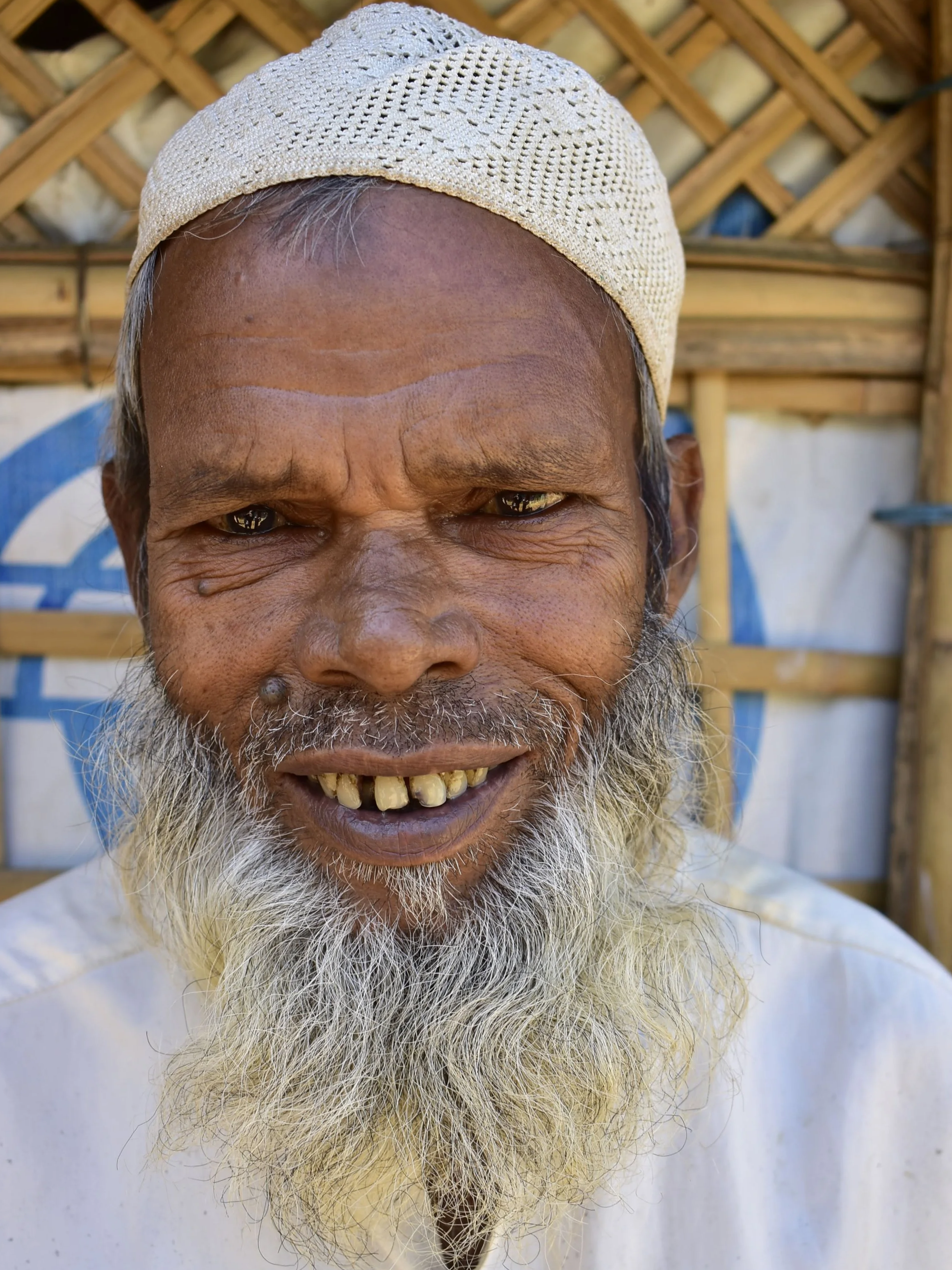 Close-up of an elderly man with a gray beard, wearing a beige knit cap, smiling, with a wooden structure in the background.