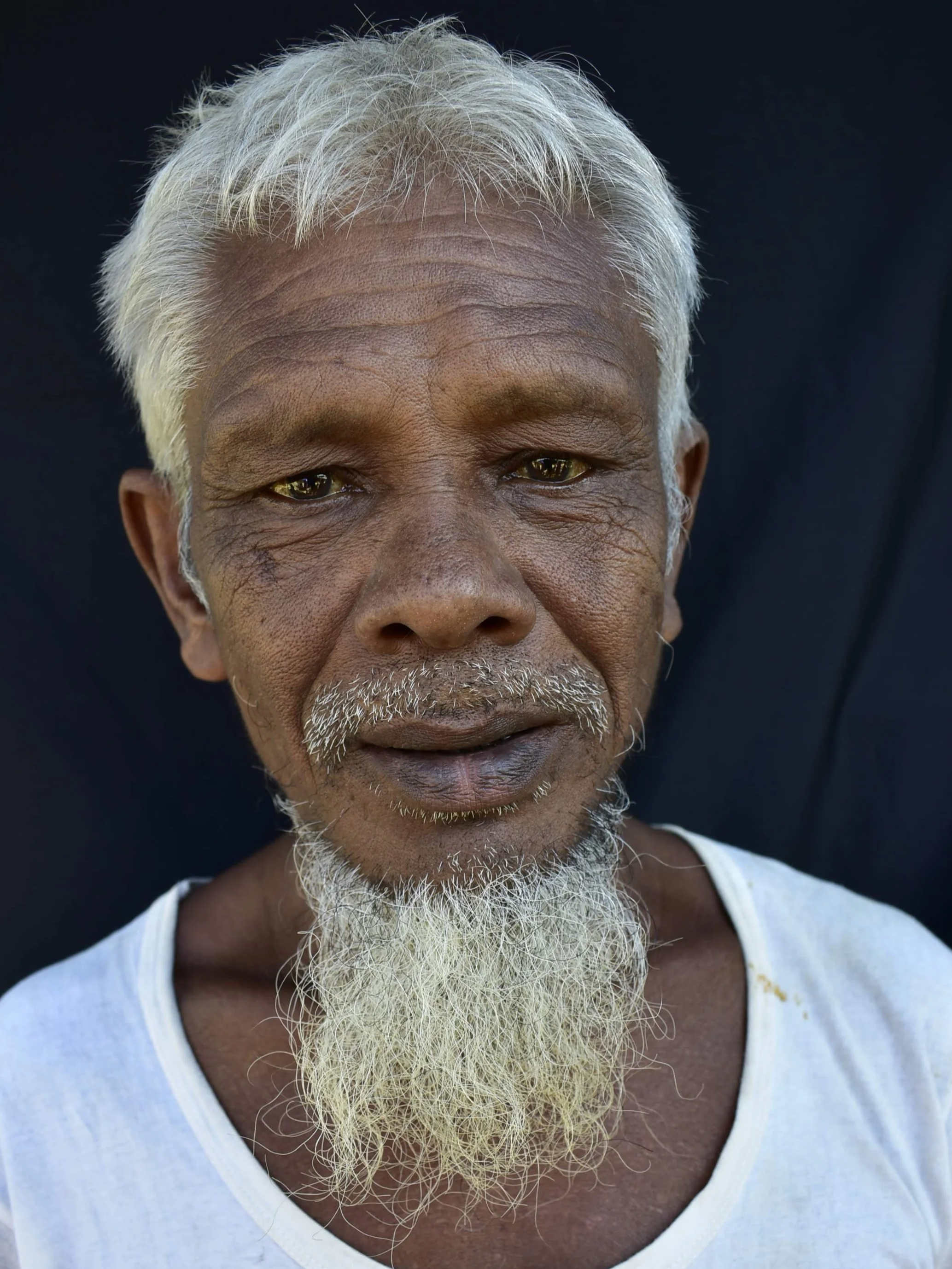 Close-up of an elderly man with a long white beard, white hair, and brown skin, wearing a white shirt, against a black background.