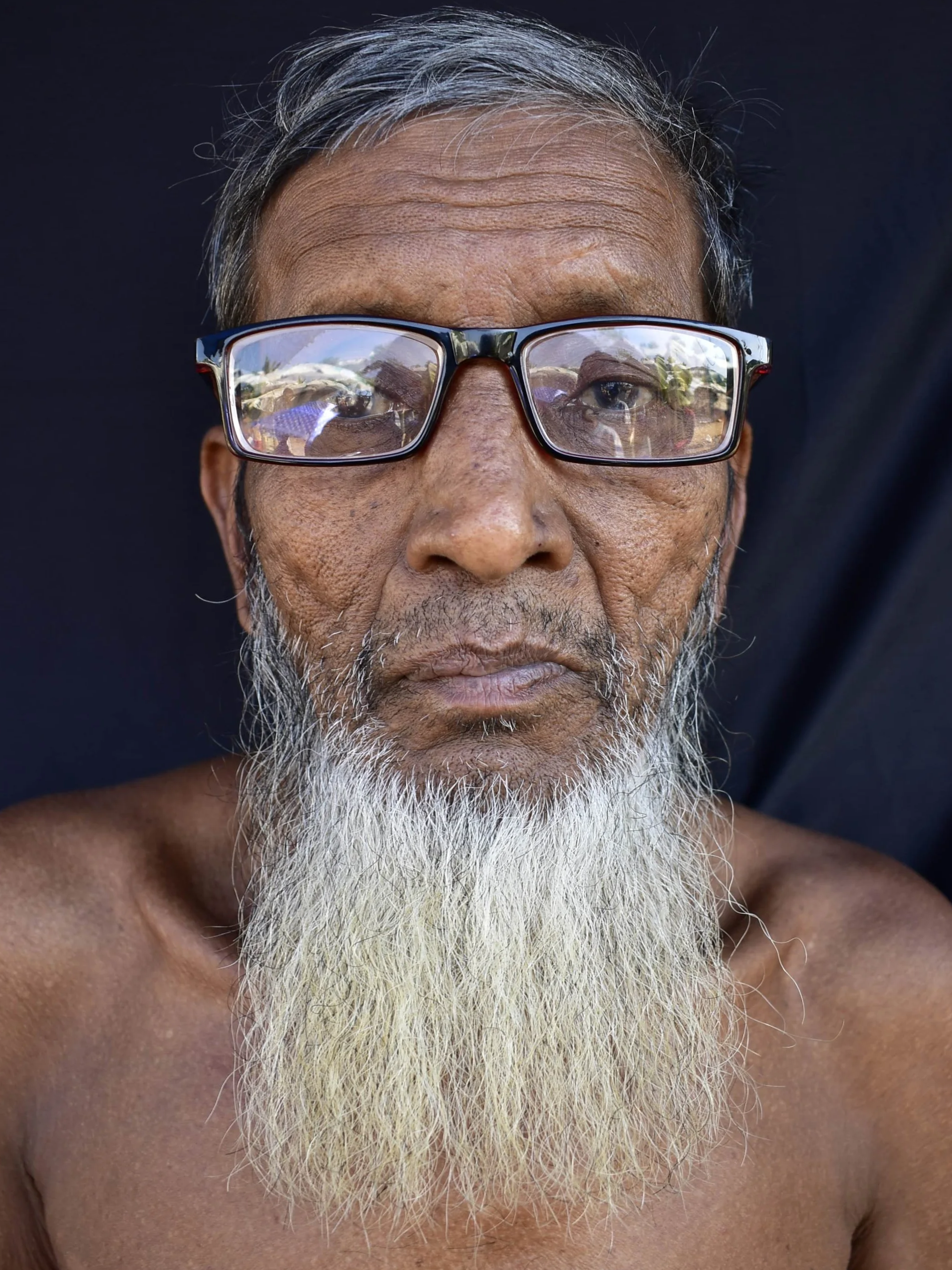 Close-up portrait of an elderly man with glasses and a long white beard, shirtless, with a dark background.