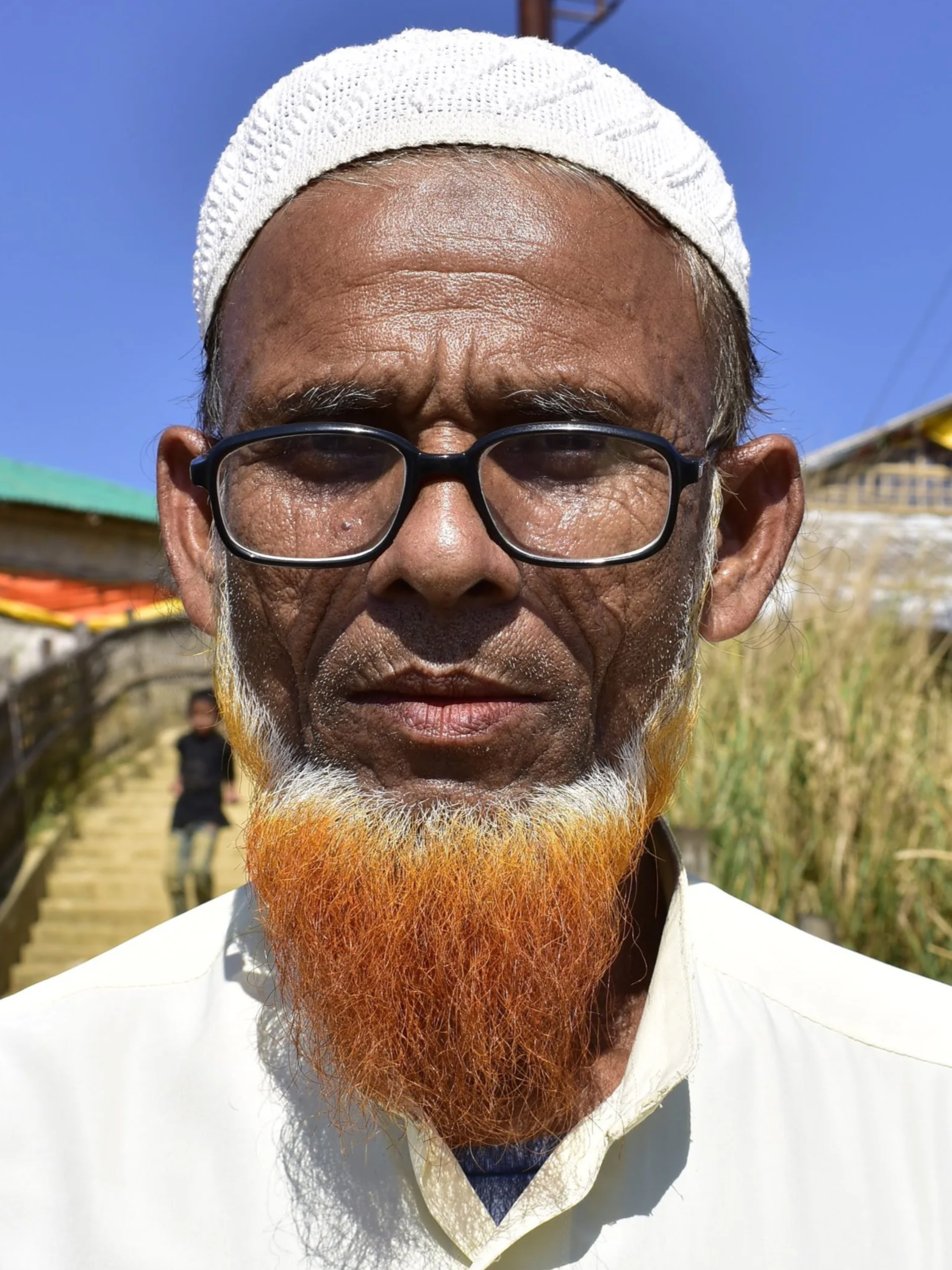 Close-up of an elderly man with glasses, a white cap, and an orange and white beard, standing outdoors against a blue sky with some trees and structures in the background.