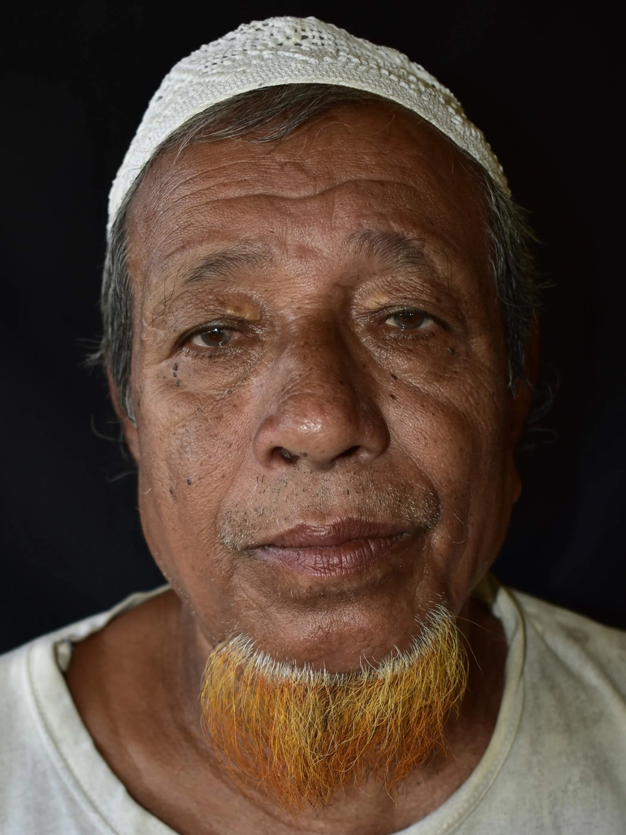 Close-up portrait of an elderly man wearing a white cap, with a yellow beard, and a neutral expression against a black background.