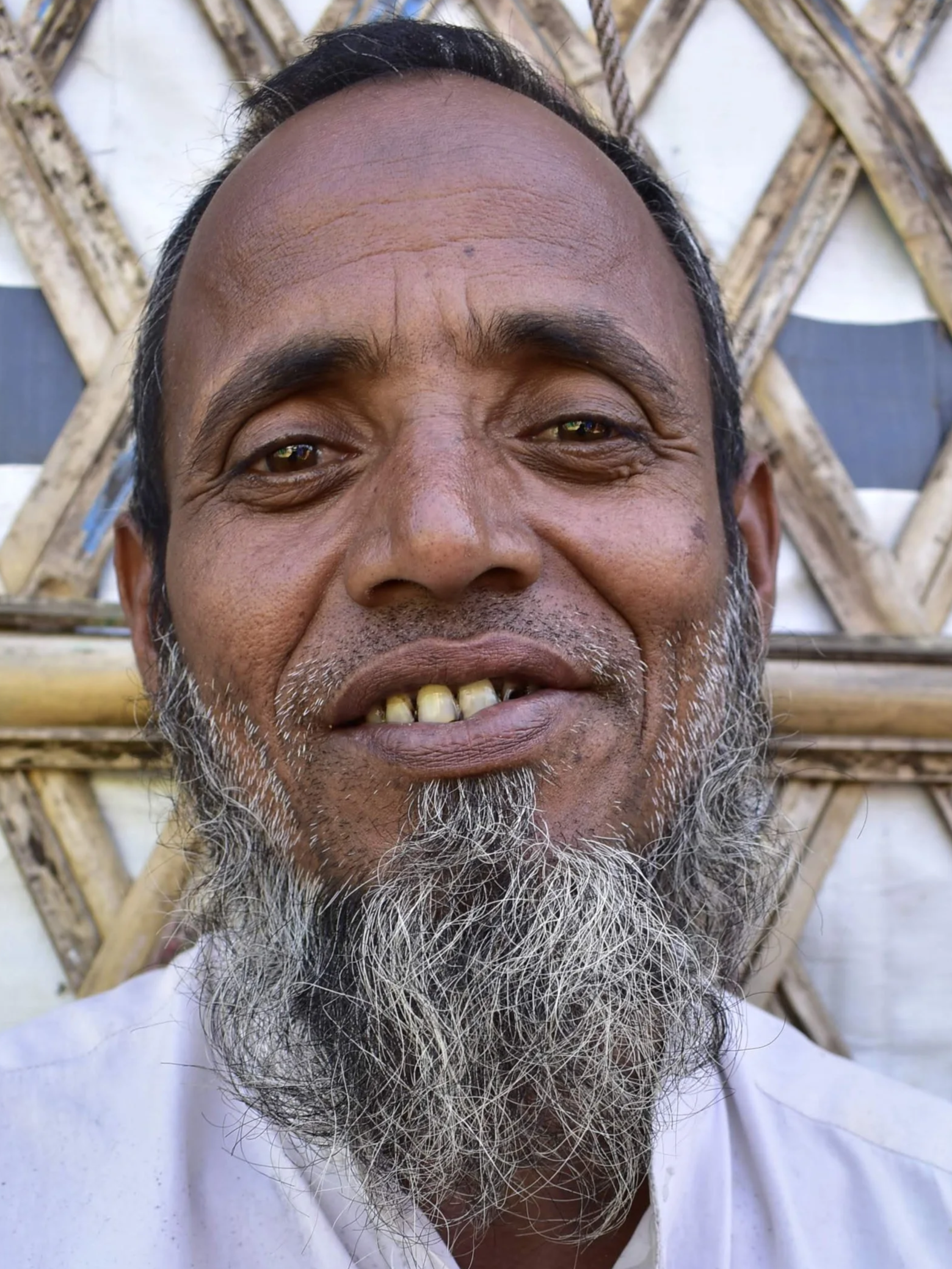 Close-up of a smiling middle-aged man with a gray beard, in front of a bamboo and white wall background.