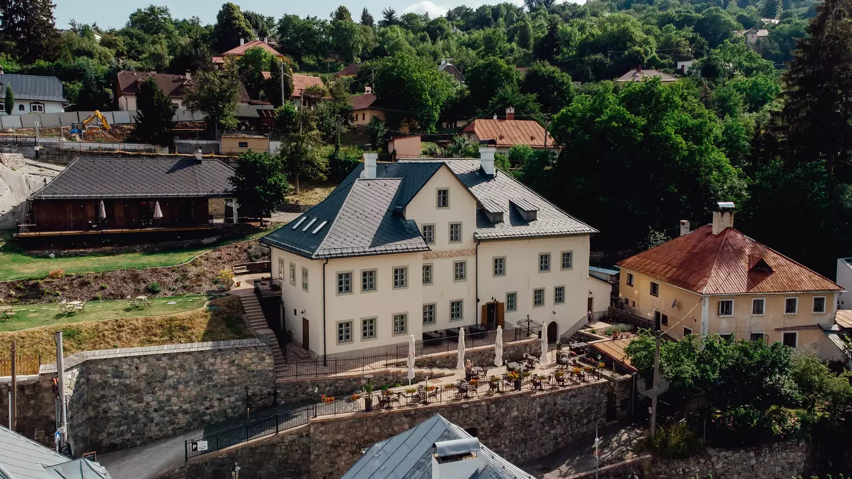 Veľký biely dom s tmavosivou strechou, viacerými oknami a terasou so slnečníkmi a záhradným nábytkom, situovaný na terasovitom kopci s kamennými múrmi. V okolí sa nachádzajú ďalšie domy, bujná zeleň a kopcovitá krajina.