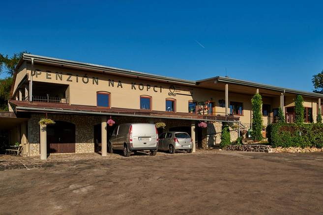 Muž stojí pri vchode do vinotéky, má na sebe ružovú zásteru a v ruke drží pohár červeného vína. Nad dverami sú vystavené fľaše vína a vonku je tabuľa s nápisom. Na stene vpravo je farebný nápis Vinoteka ViNCENTRUM. 