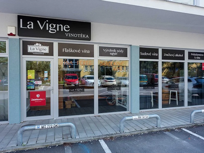 Exteriér vinotéky s názvom La Vigne, sklenenými dverami a veľkými oknami, cez ktoré je vidieť vnútornú výzdobu a zaparkované autá vonku.