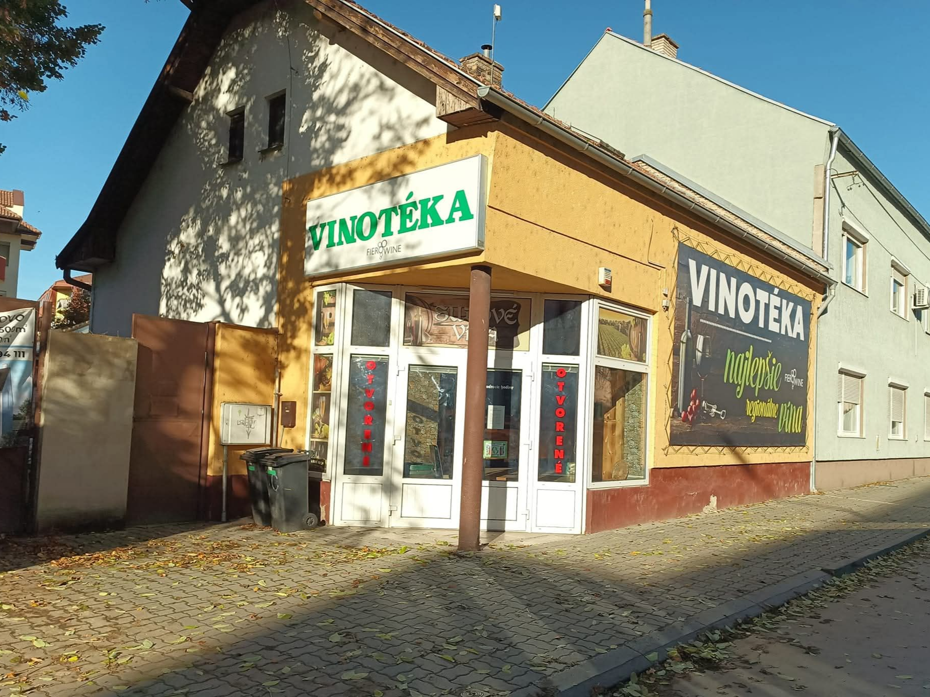 Slnečná scéna s budovou s nápisom „PEKÁREŇ“ vľavo, kostolom s červenou strechou a vežou v pozadí, cintorínom so stromami, dláždeným chodníkom a cestou zakrivenou doprava, s diaľkovými kopcami a jasnou modrou oblohou.