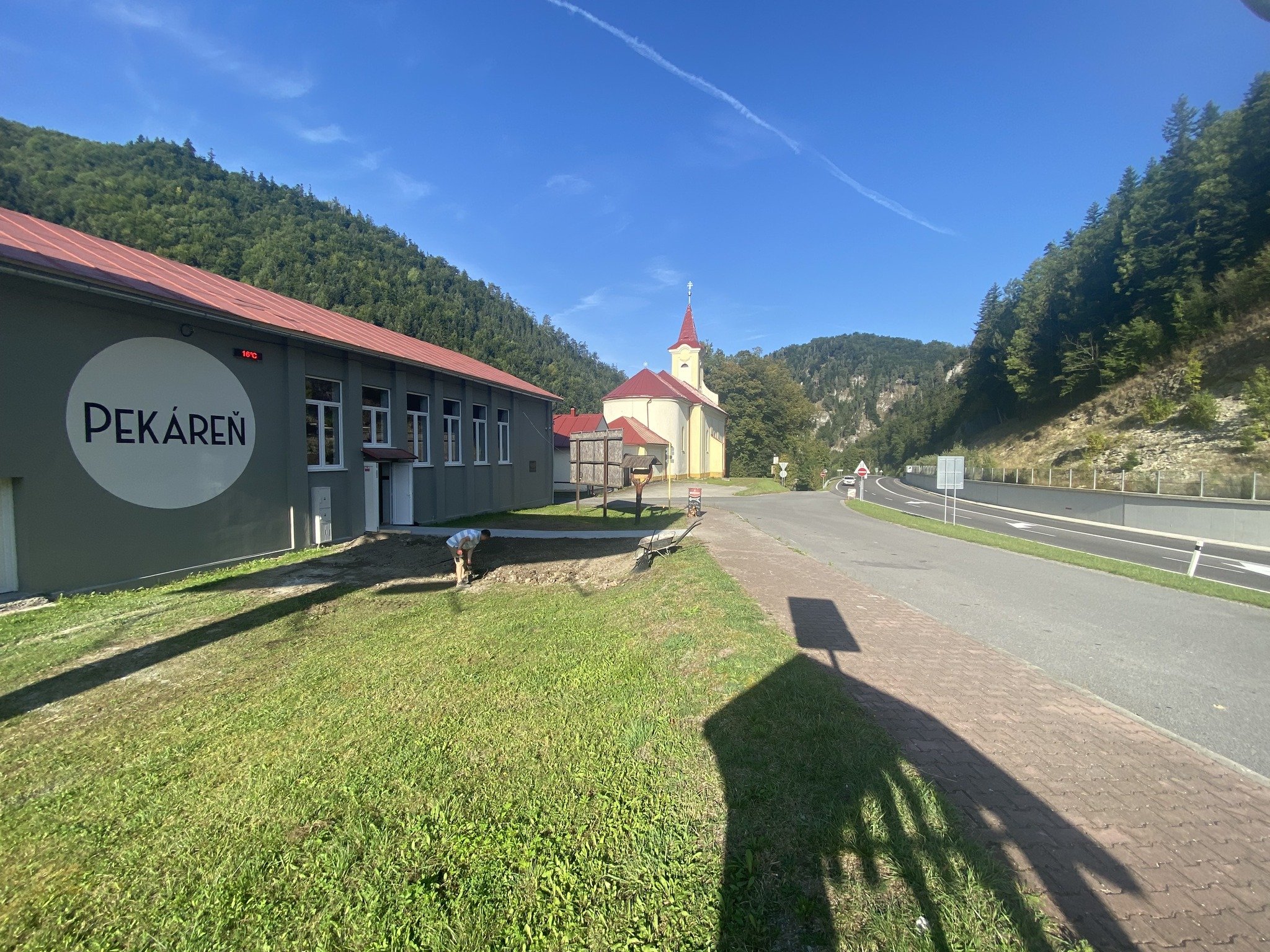 Slnečná scéna s budovou s nápisom „PEKÁREŇ“ vľavo, kostolom s červenou strechou a vežou v pozadí, cintorínom so stromami, dláždeným chodníkom a cestou zakrivenou doprava, s diaľkovými kopcami a jasnou modrou oblohou.