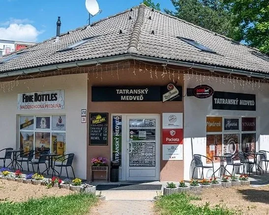 Exteriér malej budovy s nápisom „Tatranský Medveď“ a vonkajším posedením. Budova má hnedú škridlovú strechu, ružové steny a reťazové svetlá visiace z okapu. Pred budovou sú kvetinové záhony s farebnými kvetmi a obloha je čiastočne zamračená.