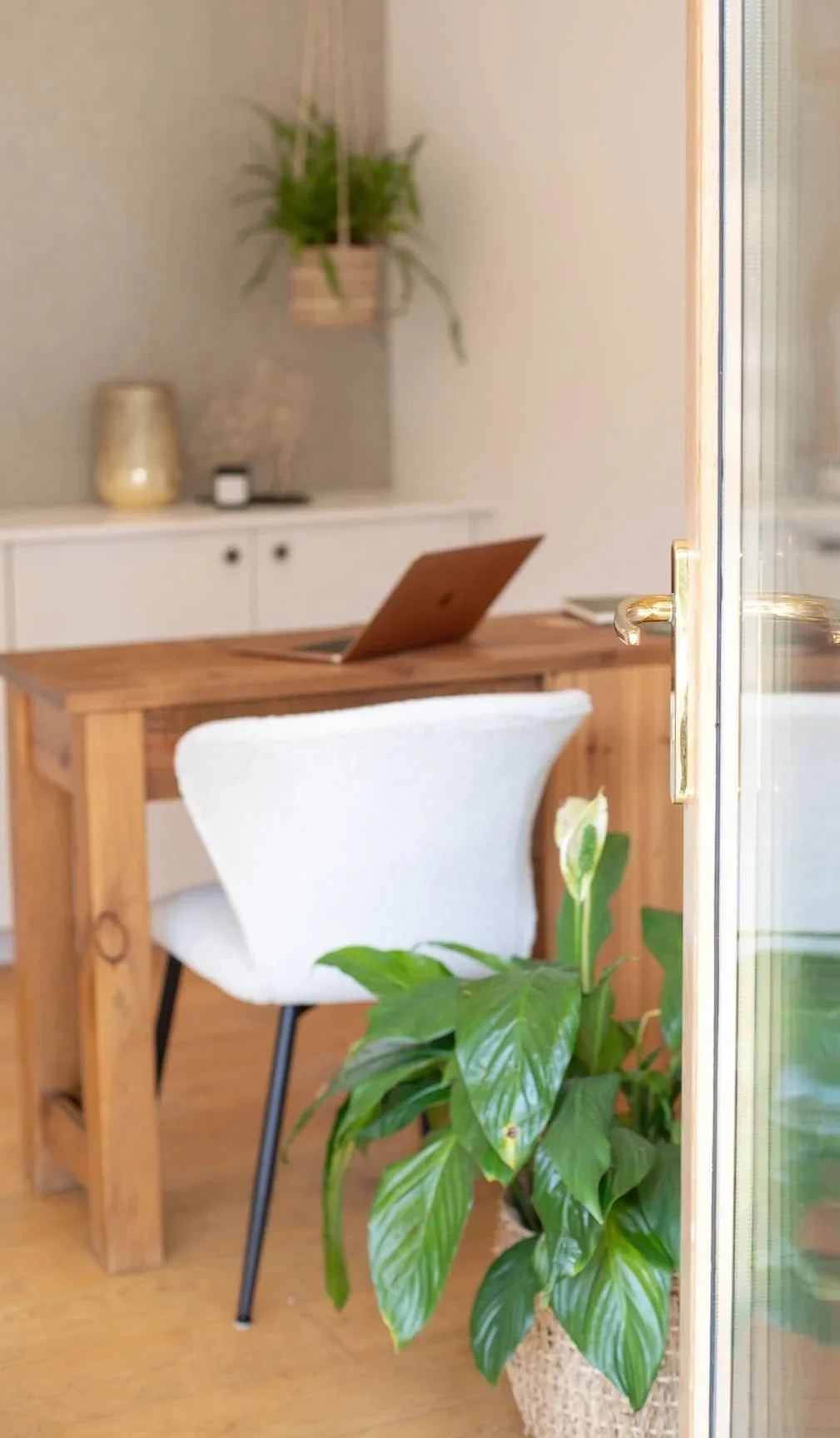 Interior of a home office with a wooden desk, a white chair, a laptop, a large potted plant, and decorative items on a white cabinet in the background.