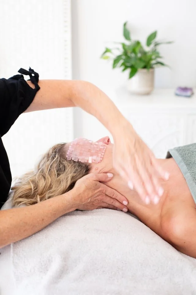 A woman receiving a massage on her neck in a spa or massage therapy setting, with a green plant in the background.