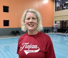 Woman with gray hair smiling, wearing a red shirt with 'Trojans' written on it, standing by an indoor swimming pool.