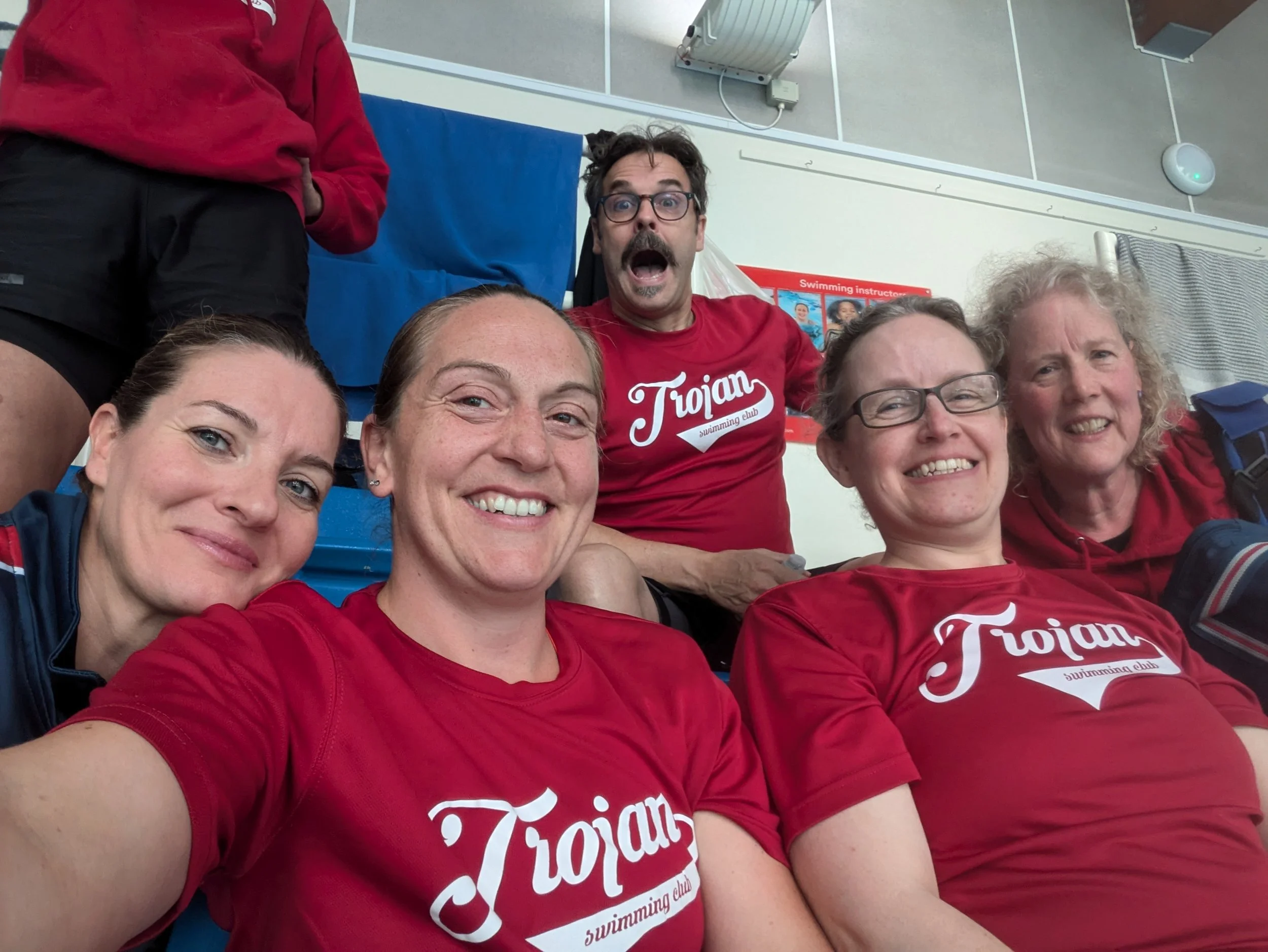 Group of five smiling women and one man taking a selfie at an indoor swimming pool, five of them wearing red T-shirts with 'Trojan swimming club' printed on them. One woman is wearing a navy jacket, and one man in a red T-shirt is making a surprised 