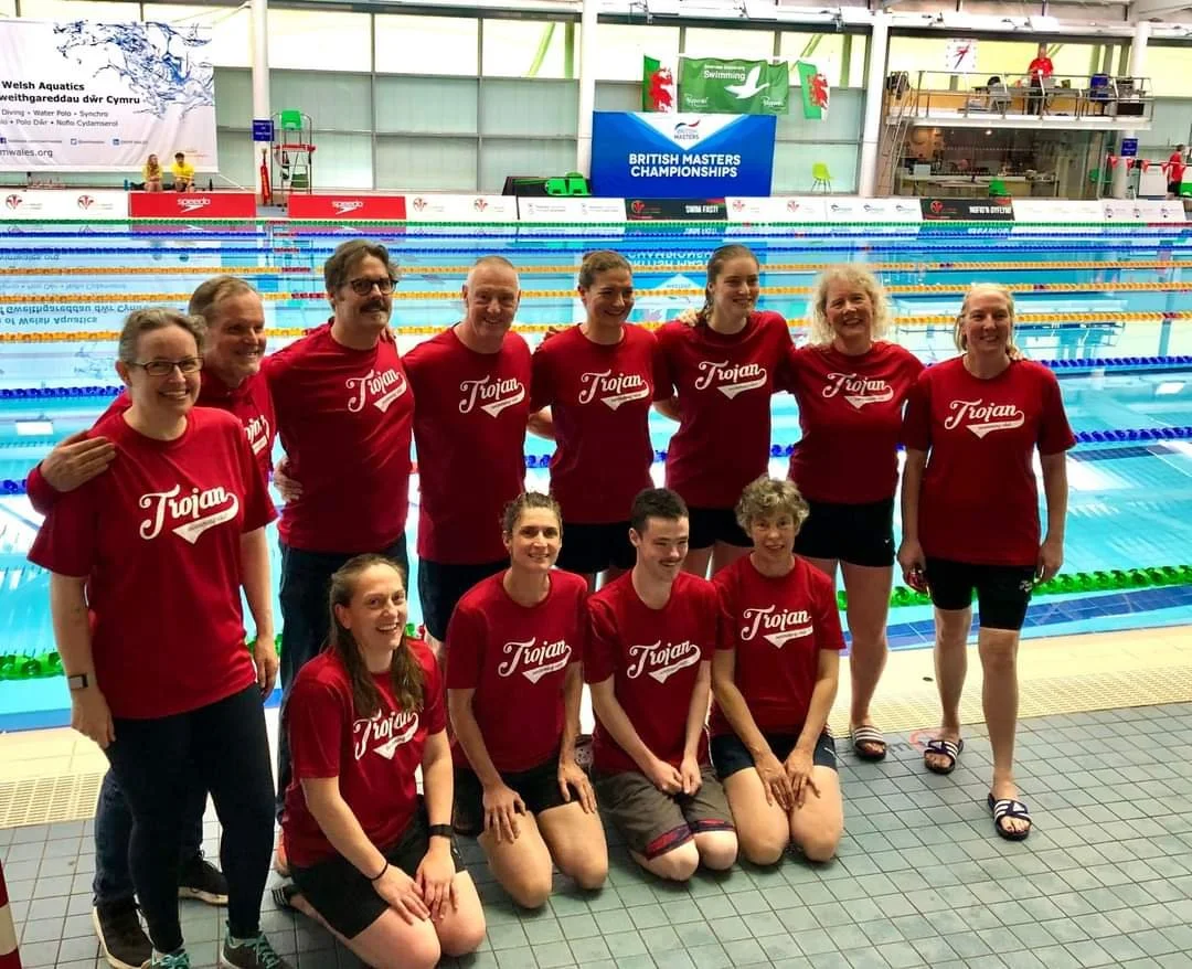 A group of eleven swimmers, all wearing matching red T-shirts with "Trojan" printed on them, posing for a team photo at an indoor swimming pool during the British Masters Championships.
