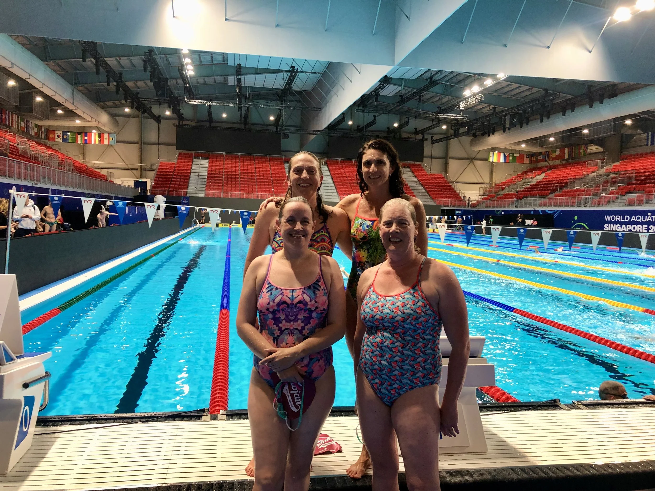 Four women in swimsuits standing together at the edge of an indoor swimming pool during a swim meet.