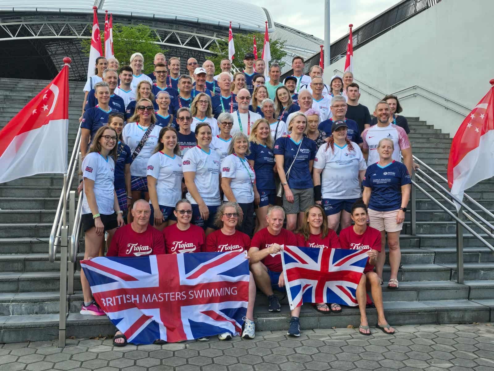 Group photo of people holding British and Singapore flags on stairs outside a stadium, celebrating at a swimming event.