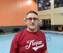 A man wearing glasses and a red Trojans t-shirt standing at the edge of an indoor swimming pool.