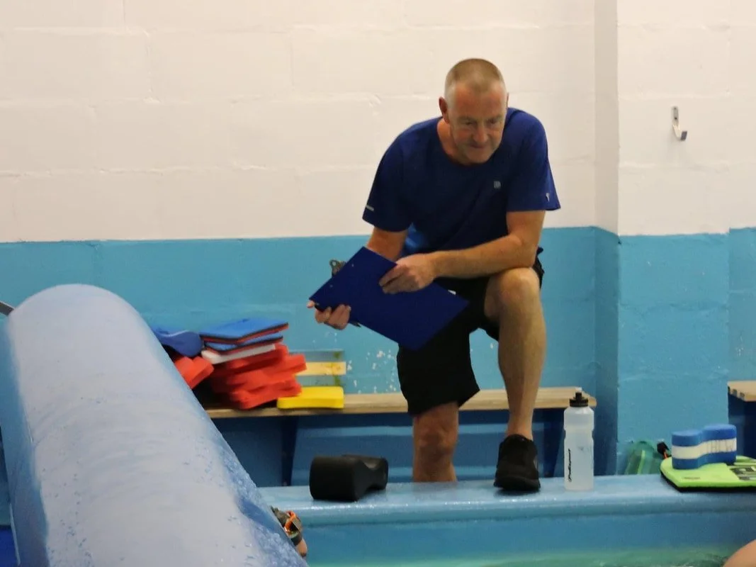 Man in athletic clothing standing on one leg on a pool edge, holding a clipboard, with stacks of floatation belts nearby.