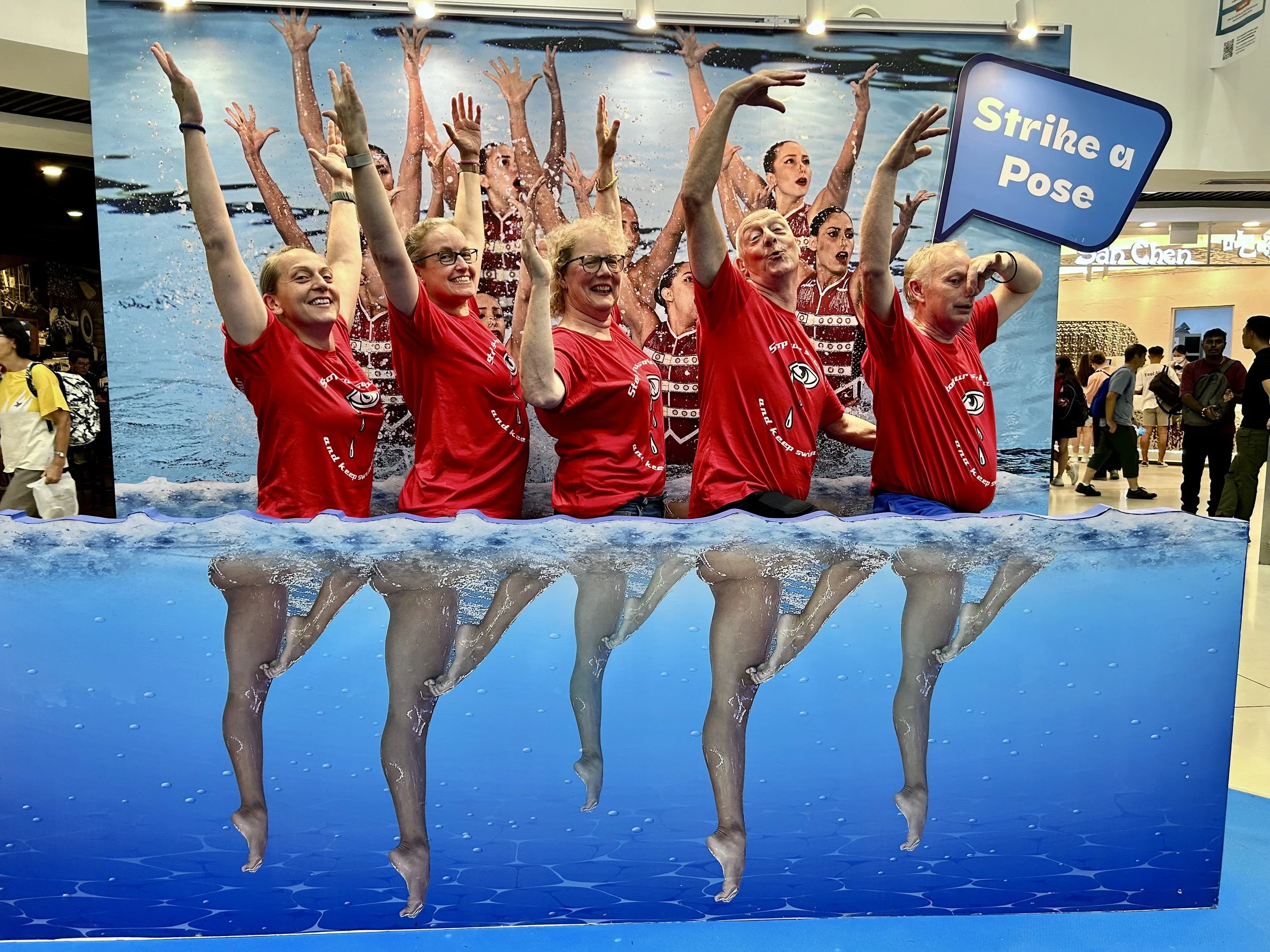 A display with a backdrop of women swimming and water splashing, showing six women in red shirts standing in a water-themed cutout, raising their arms, with a speech bubble saying 'Strike a Pose'.