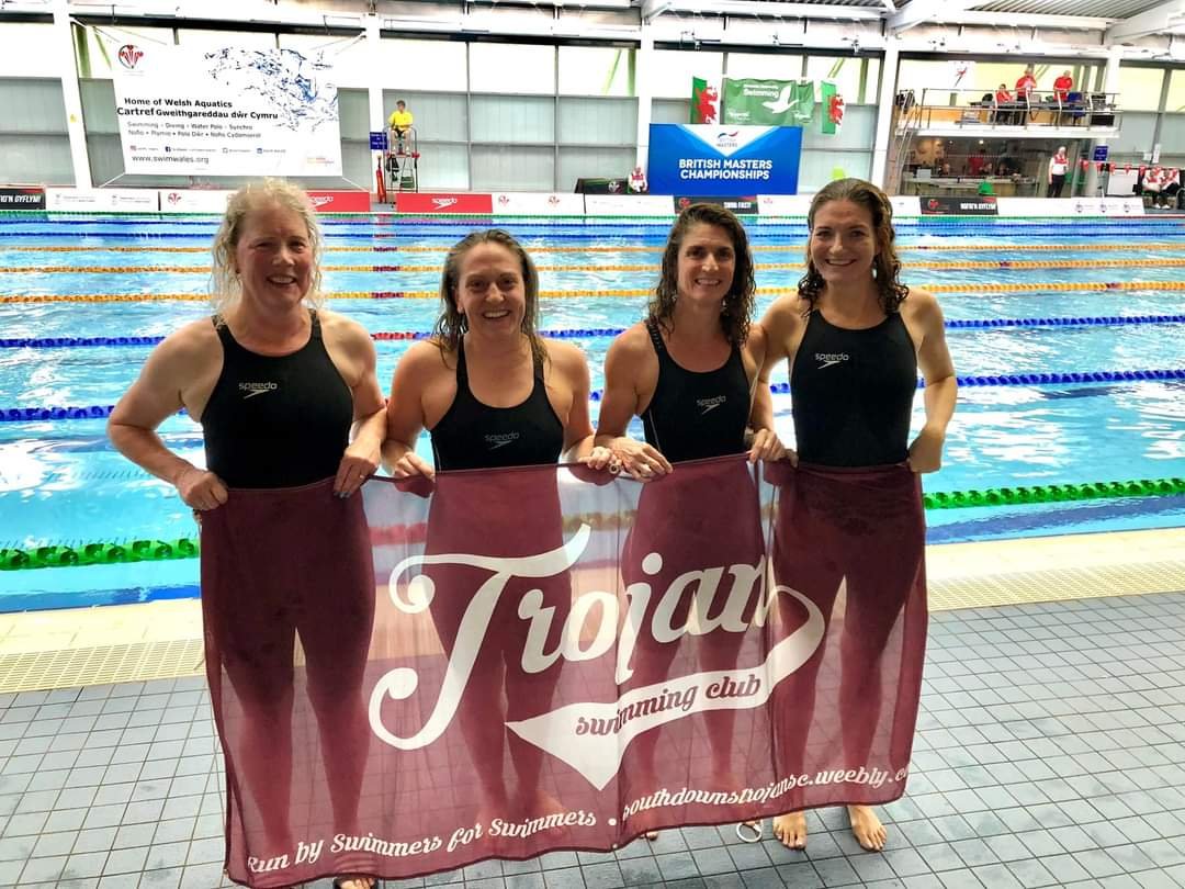 Four female swimmers in black swimsuits holding a maroon banner that reads 'Trojan swimming club,' standing poolside at an indoor swimming pool during a competition. The pool has multiple lanes with floating lane dividers, and there are signs and ban