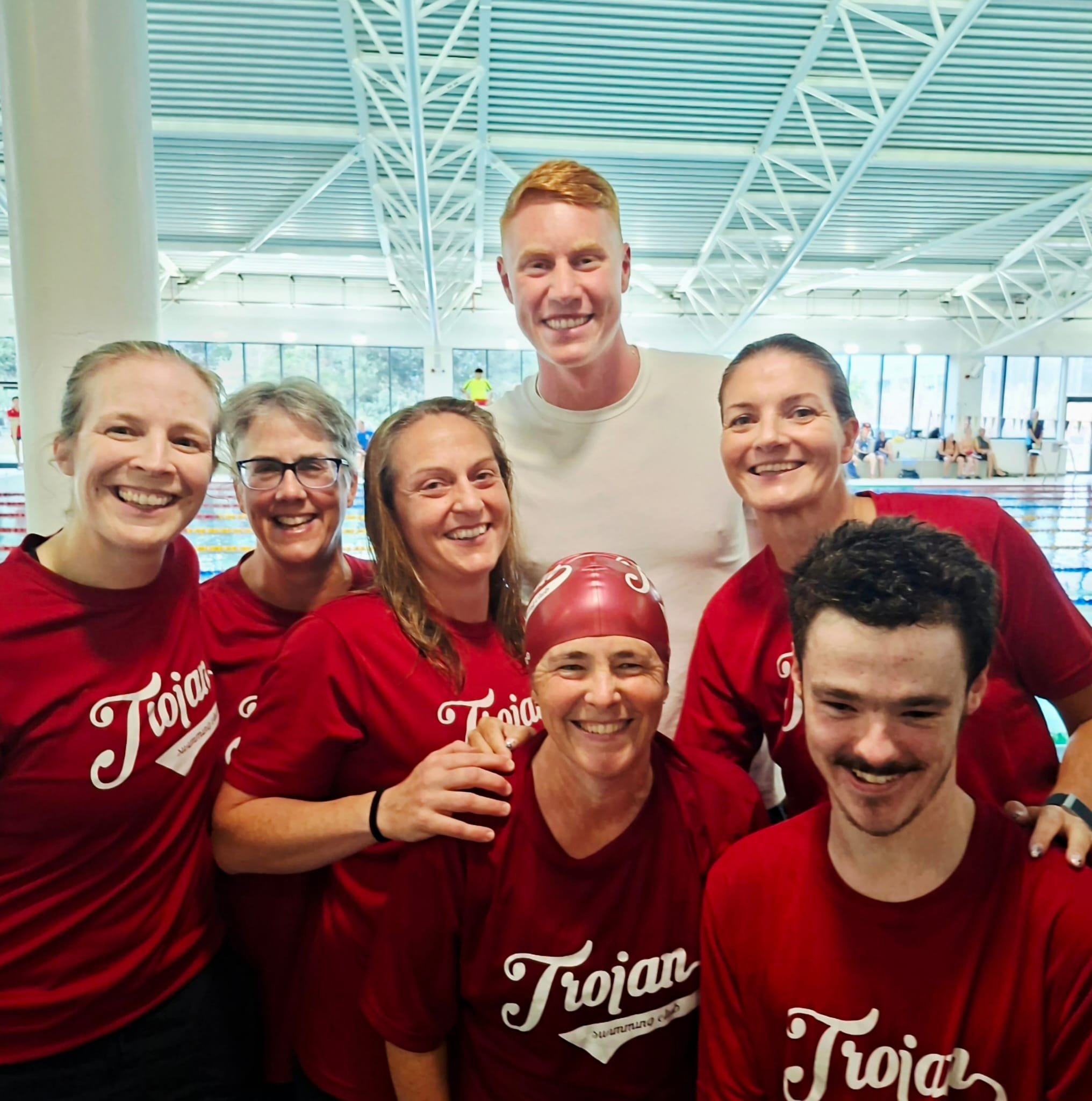 Group of six swimmers in red Trojan shirts, some wearing swim caps, posing together inside an indoor swimming pool.