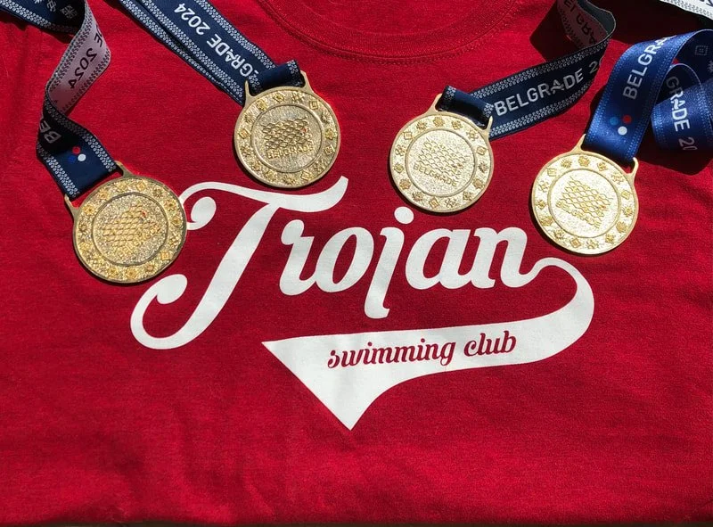 Four gold medals with blue and black ribbons, placed on a red Troian swimming club shirt.