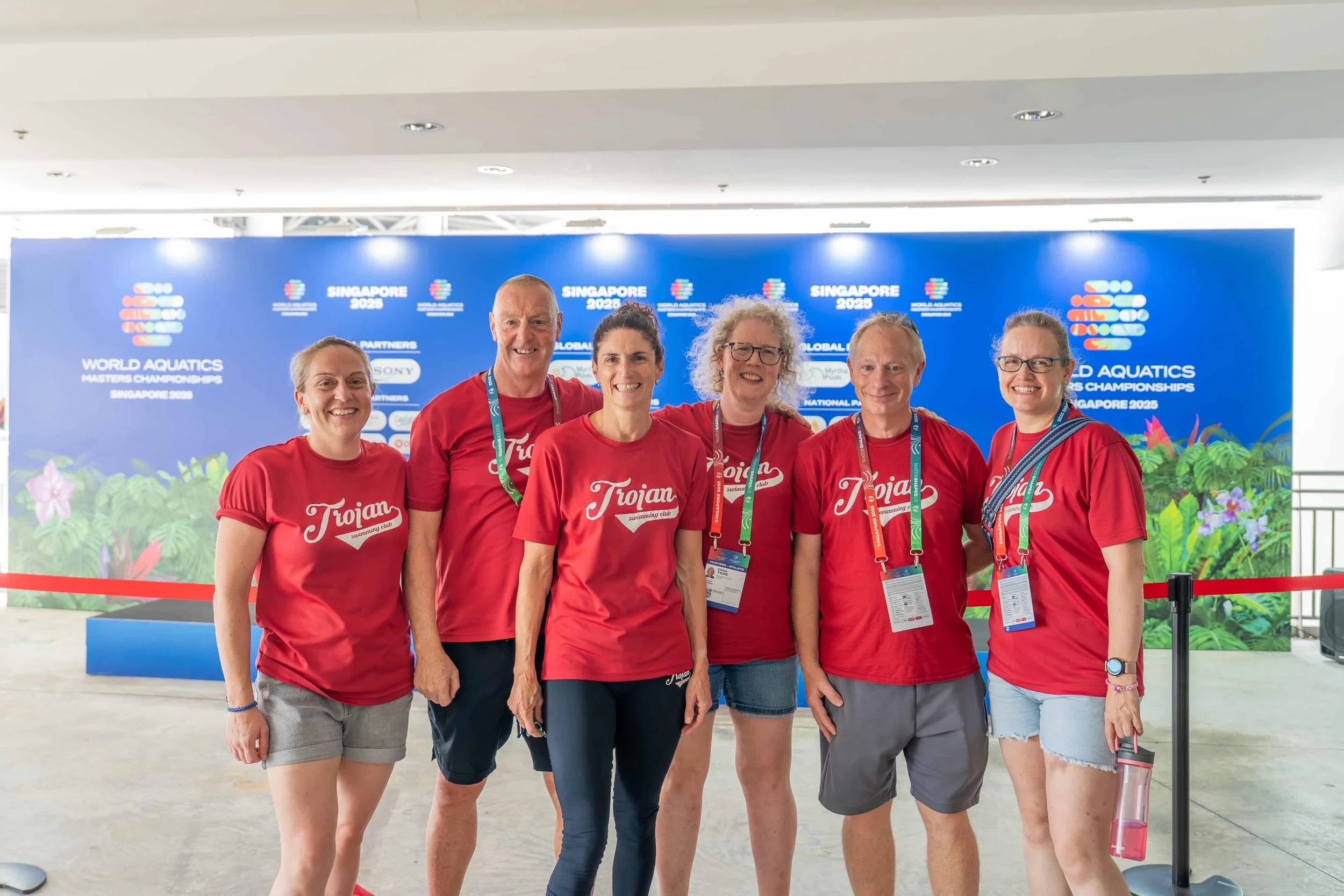 Group of six smiling people wearing red 'Trojan' shirts at the World Aquatics Championships in Singapore 2023, standing in front of a blue event backdrop.