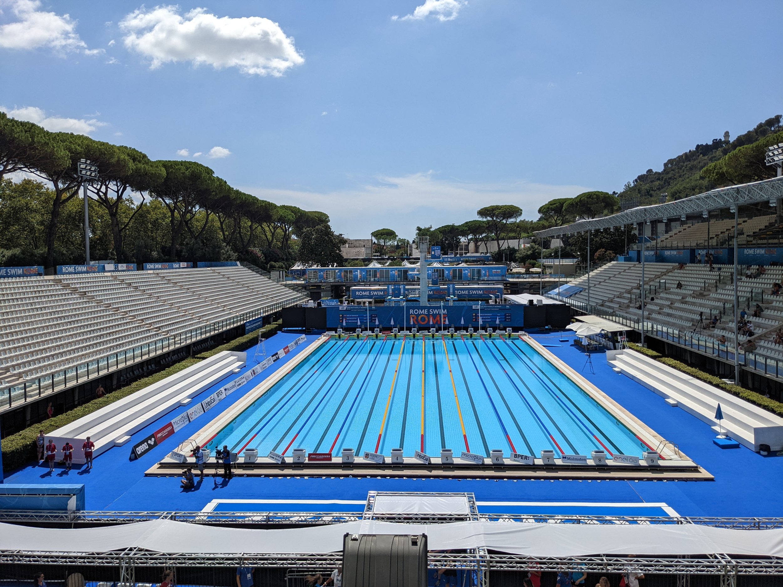 Olympic-sized swimming pool at an outdoor venue, with seating on both sides, surrounded by trees, under a blue sky.