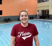 A woman in a red Trojans t-shirt standing by a swimming pool, smiling.