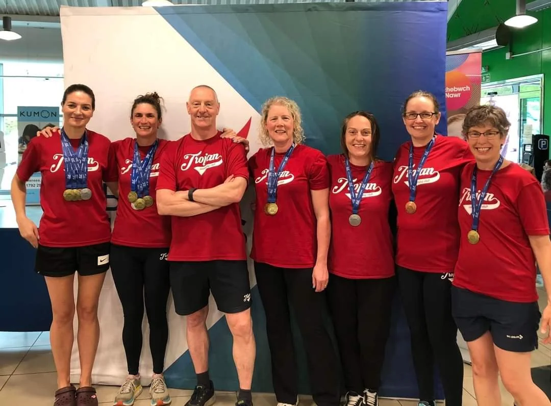 Group of seven people, six women and one man, wearing matching red shirts with 'Trojan' written on them, standing together indoors, each wearing medals around their necks, smiling and posing for the photo.