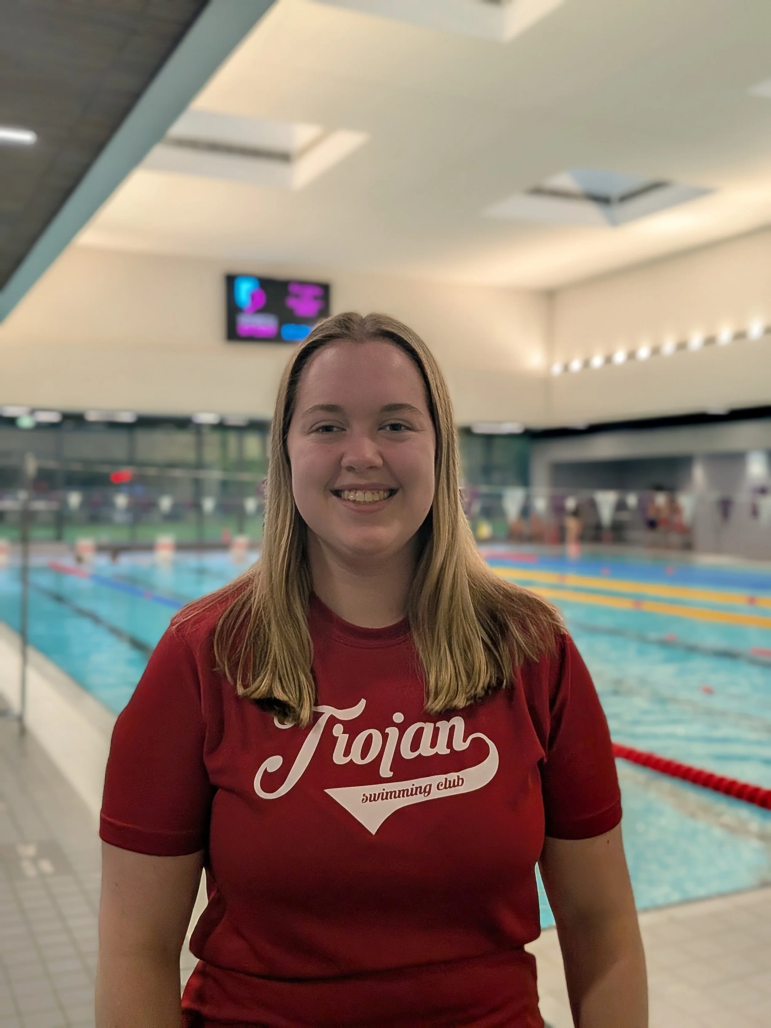 A smiling young woman with long blond hair standing inside a swimming pool facility, wearing a red T-shirt that says 'Trojans Swimming Club'.