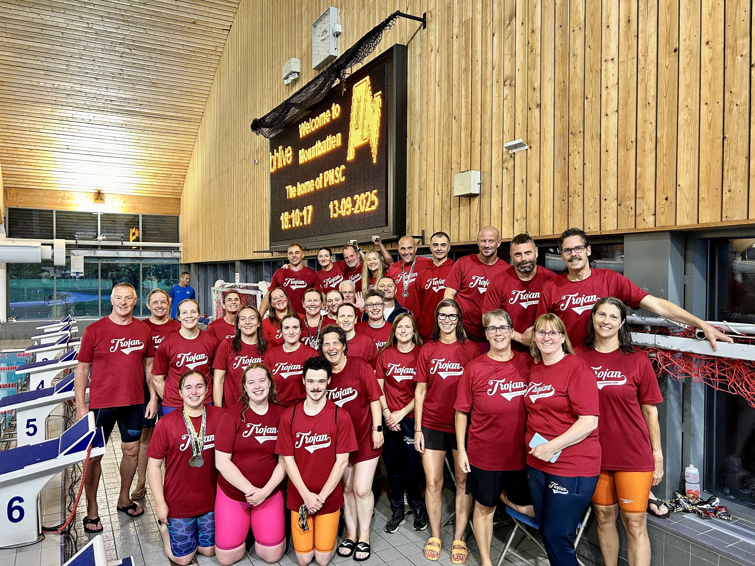 Group of people in swimming attire and red T-shirts with 'Trojan' printed on them, standing in an indoor swimming pool area, celebrating after a swim meet, with a scoreboard displaying meet details in the background.