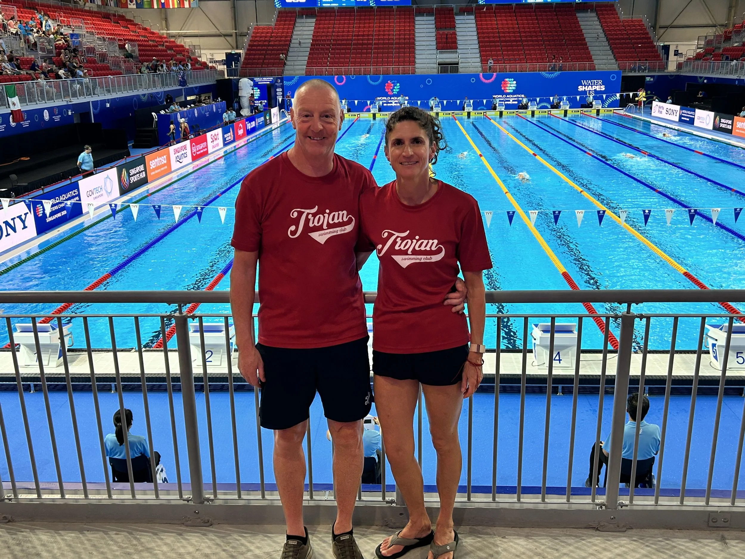 A man and woman in red 'Trojan Swimming Club' T-shirts standing together at an indoor swimming pool. The pool has multiple swimming lanes, and there are spectators and officials in the background.
