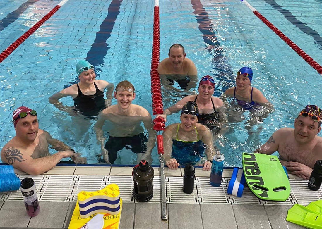 A group of nine people in a swimming pool, smiling and posing for a photo, with swim gear and water bottles on the pool deck in front of them.