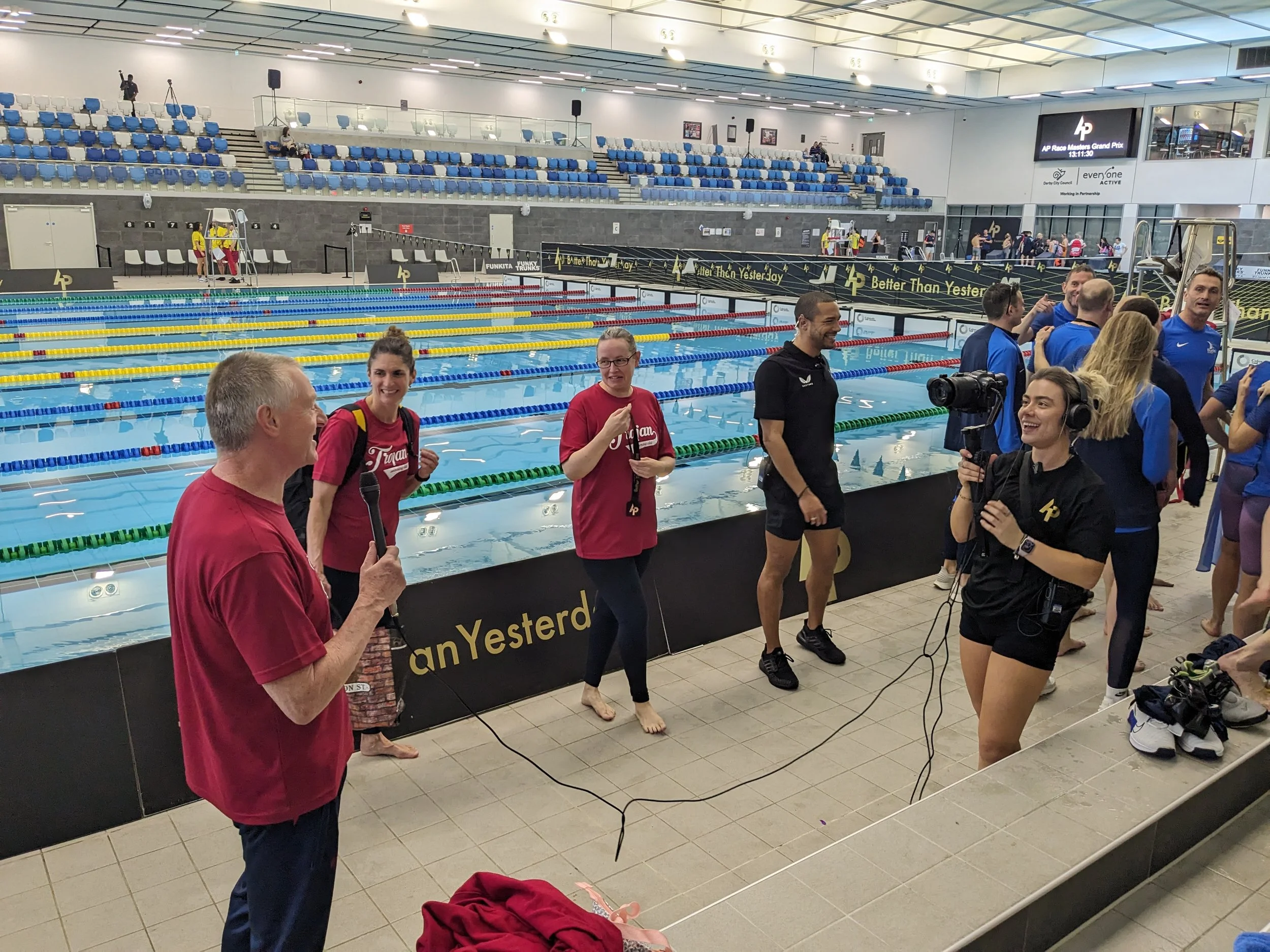 Swimmers, coaches, and a videographer at an indoor swimming pool during a media interview or event, with swimming lanes in the background and seating areas above.