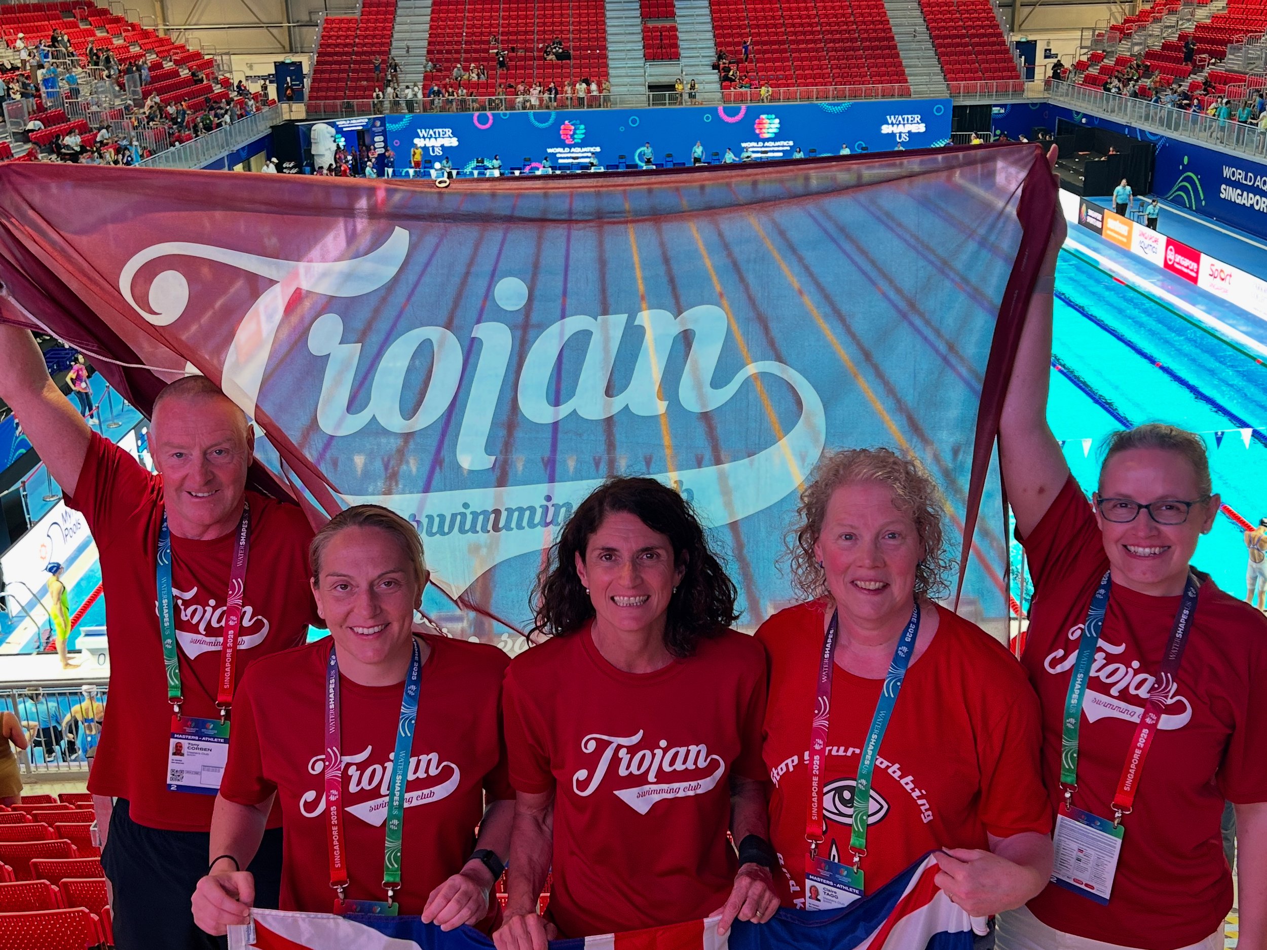 Group of five swimmers wearing red T-shirts with 'Trojan swimming club' logo, holding a flag, inside an indoor swimming pool arena at a swim meet, with red seats and swimming lanes in the background.