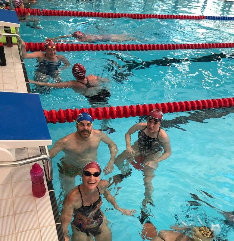 Swimmers at an indoor swimming pool, wearing swim caps and goggles, standing next to the pool edge and smiling at the camera.