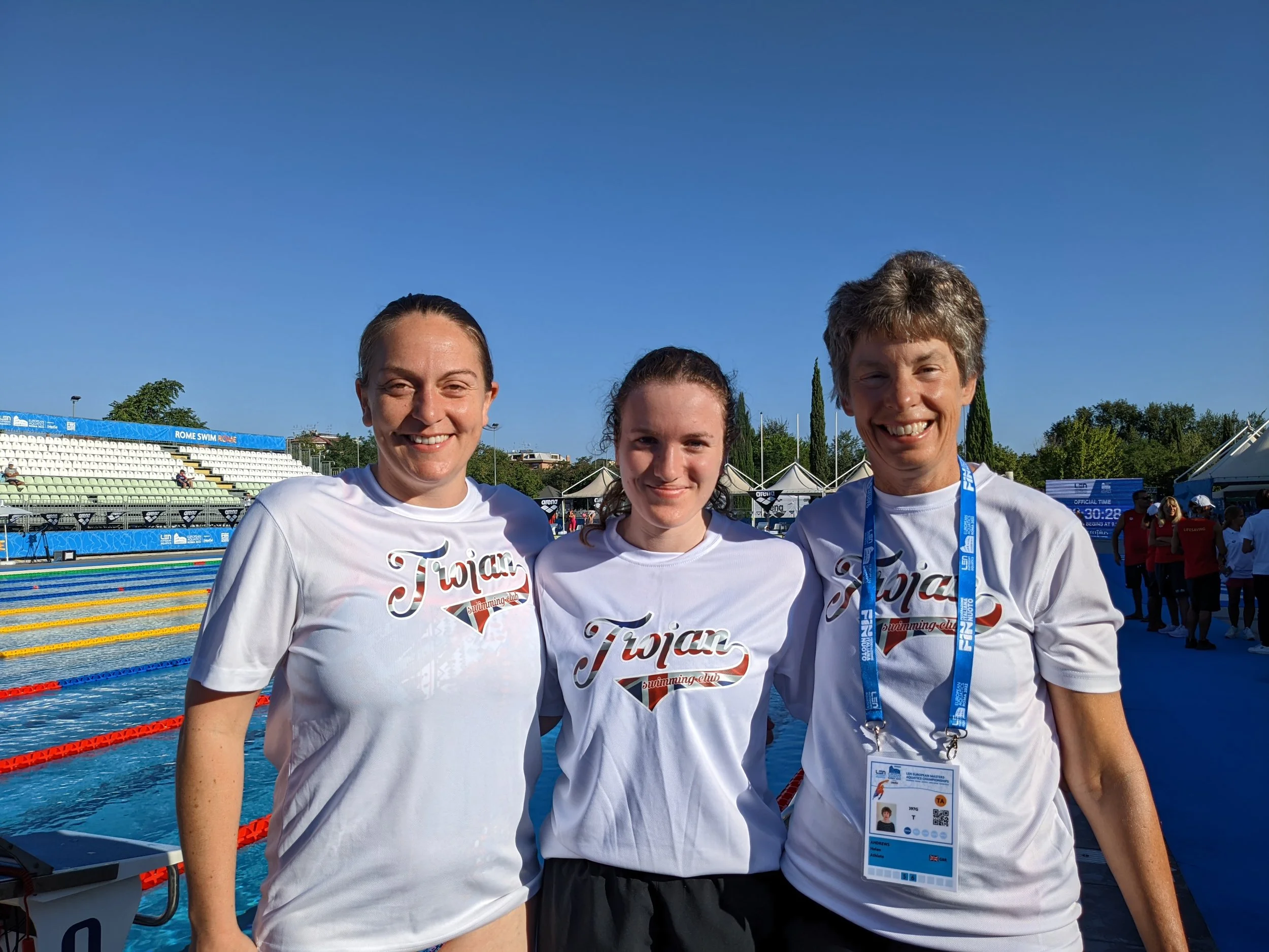 Three swimmers in white T-shirts with 'Tijan' printed on them, smiling and posing together by the poolside during a swimming event.