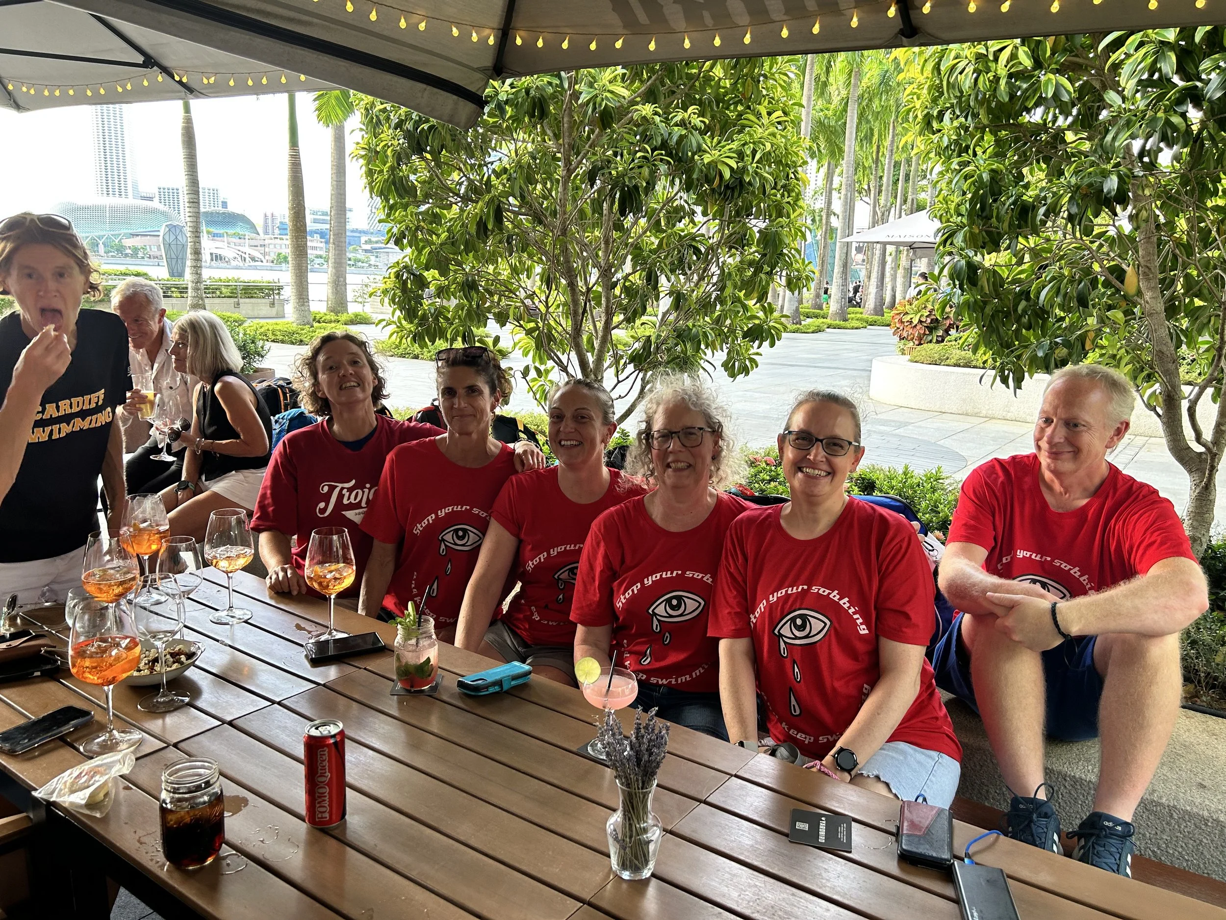 A group of six smiling people sitting at a wooden outdoor table, some wearing red shirts with a logo, with drinks and snacks, under a shaded canopy with a cityscape and greenery in the background.