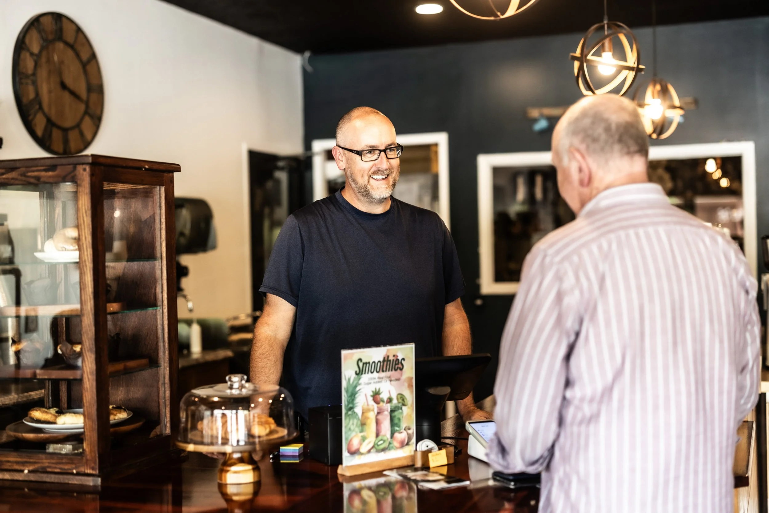 A man with glasses and a beard smiling at a customer in a cafe counter with pastries, a smoothie menu, and a cash register.