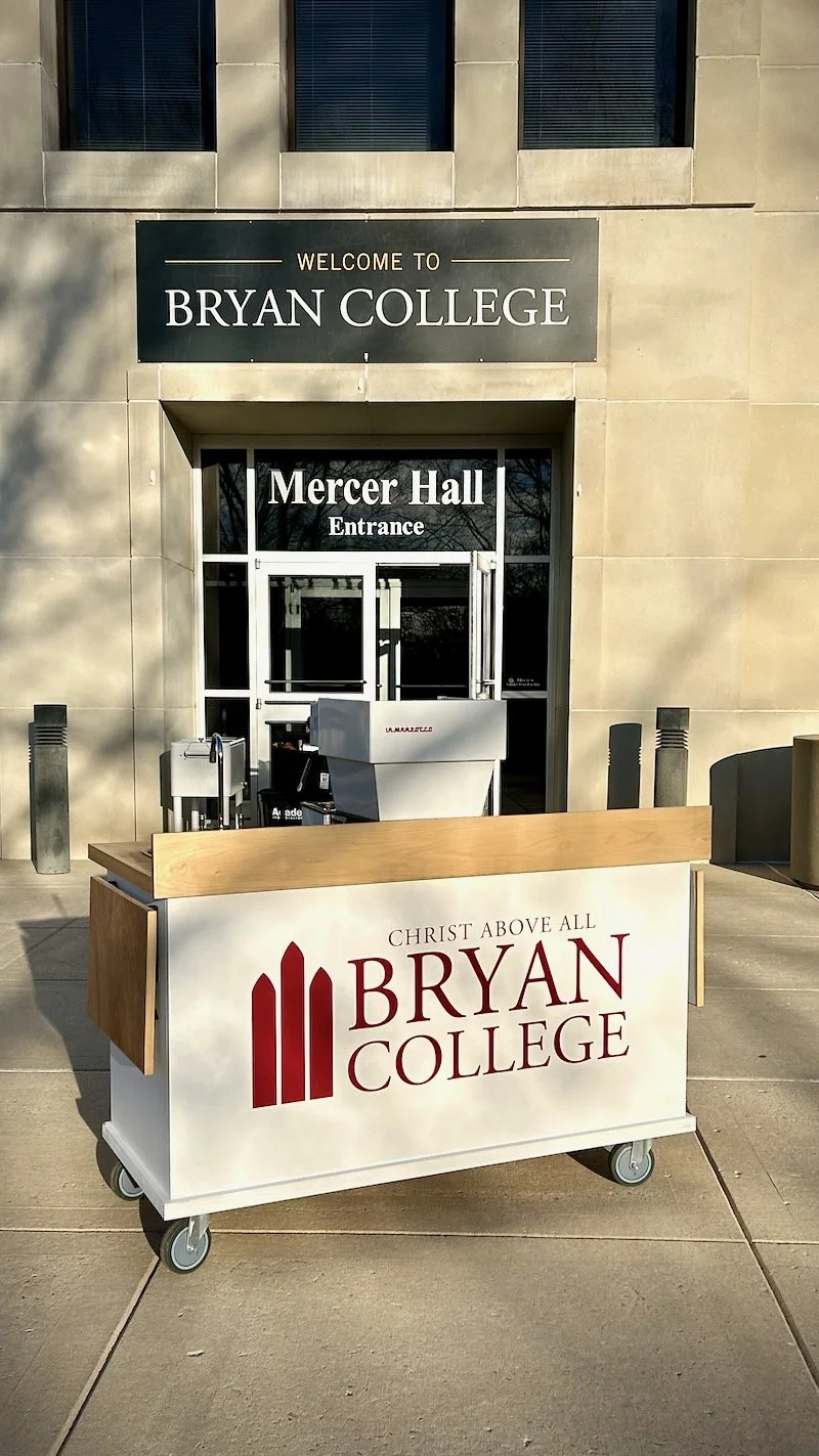 A college entrance sign for Bryan College with a welcome sign, a coffee cart ready to roll in, and a building entrance labeled Mercer Hall.