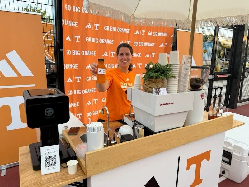 A woman in a bright orange T-shirt is standing behind a coffee stand, holding up a bottle with a logo. The stand is decorated with orange and white branding for the University of Tennessee, including the messages "GO BIG ORANGE." There are cups, a co