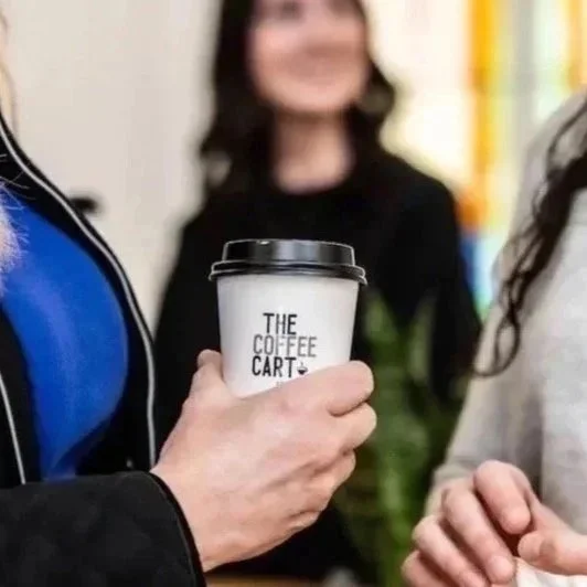 Person holding a coffee cup with the text 'The Coffee Cart' printed on it, while other people are present in the background.