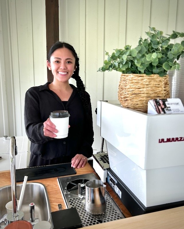 A woman with dark hair smiling and holding a cup of coffee, standing behind The Coffee Cart next to a coffee machine catering a wedding at Howe Farms venue near Chattanooga Tennessee. 