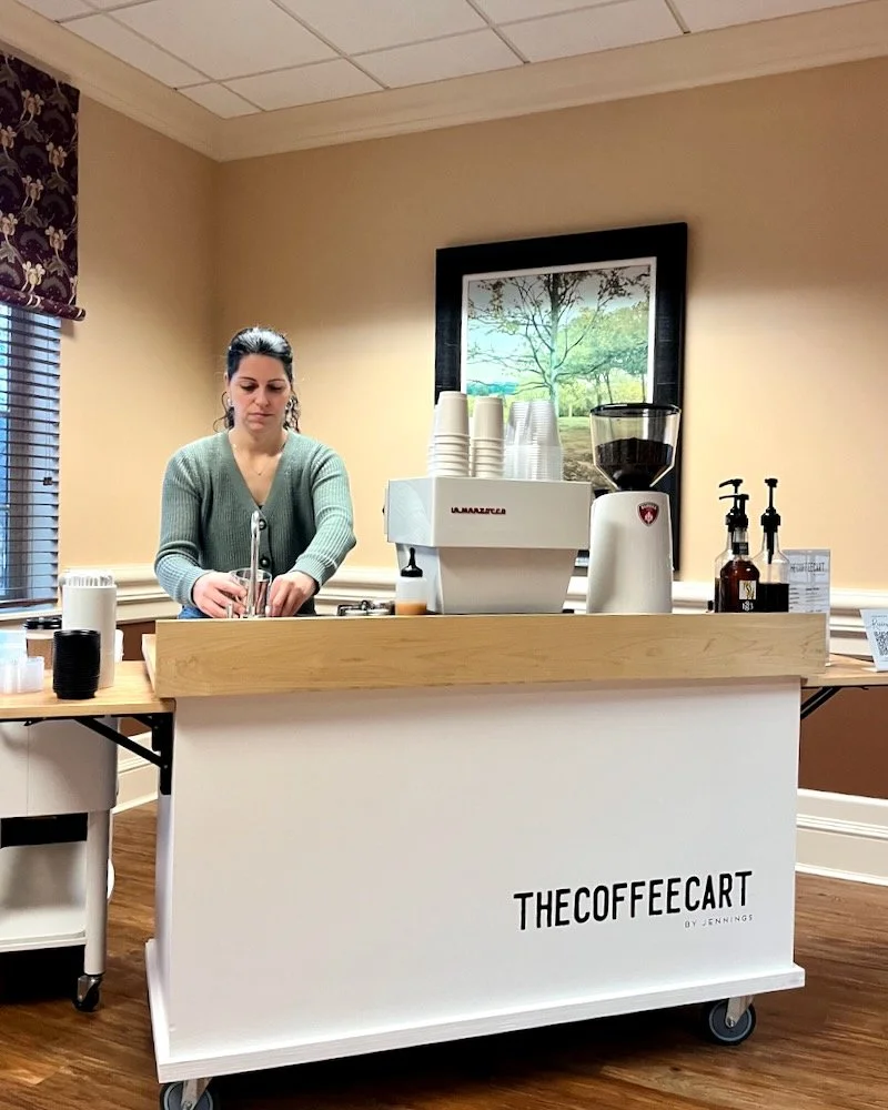 Woman preparing coffee behind a mobile coffee cart labeled 'The Coffee Cart by Jennings' in a room with beige walls, window with blinds, and a framed picture of a tree outside.