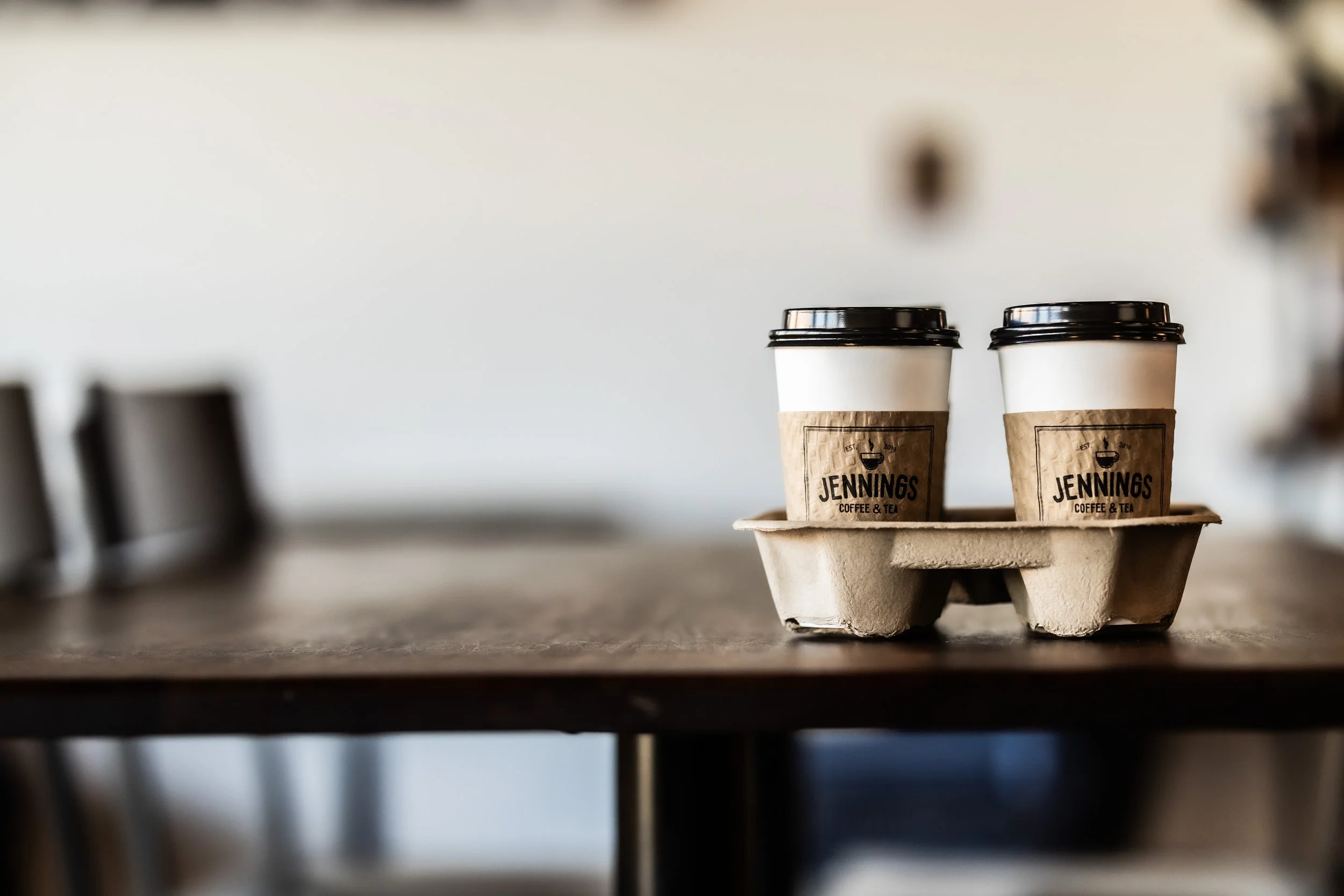 Two disposable coffee cups with black lids on a tray, placed on a wooden table inside a coffee shop.