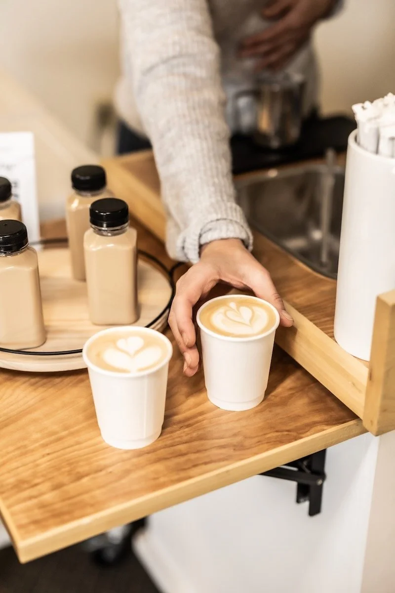 A person serving two cups of coffee with latte art from a coffee cart in a event setting, with bottled lattes on a wooden tray.