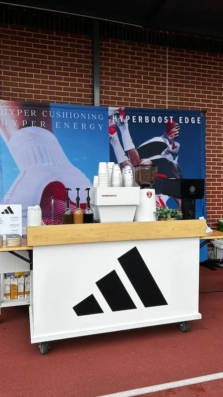 A full coffee cart setup with a custom cart with the Adidas logo, featuring coffee cups, a coffee grinder, syrup bottles, and iced coffee containers, set against a sports-themed backdrop with an athlete in a dynamic pose.